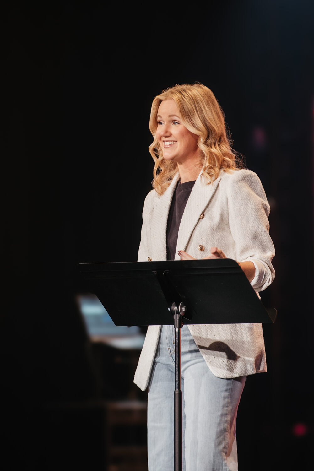 Luann Riley preaching at a lectern in a white jacket, smiling mid-message